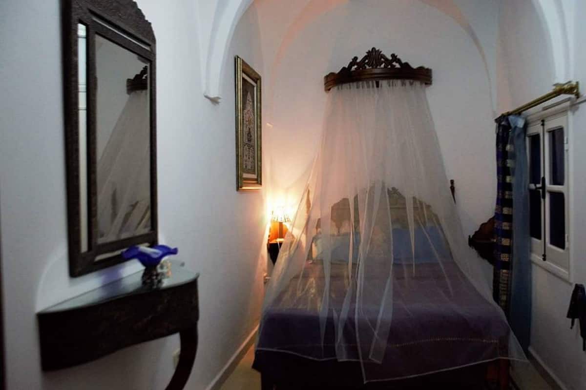 A narrow room with a bed under a canopy, a bedside table with a lamp, and a window with curtains.