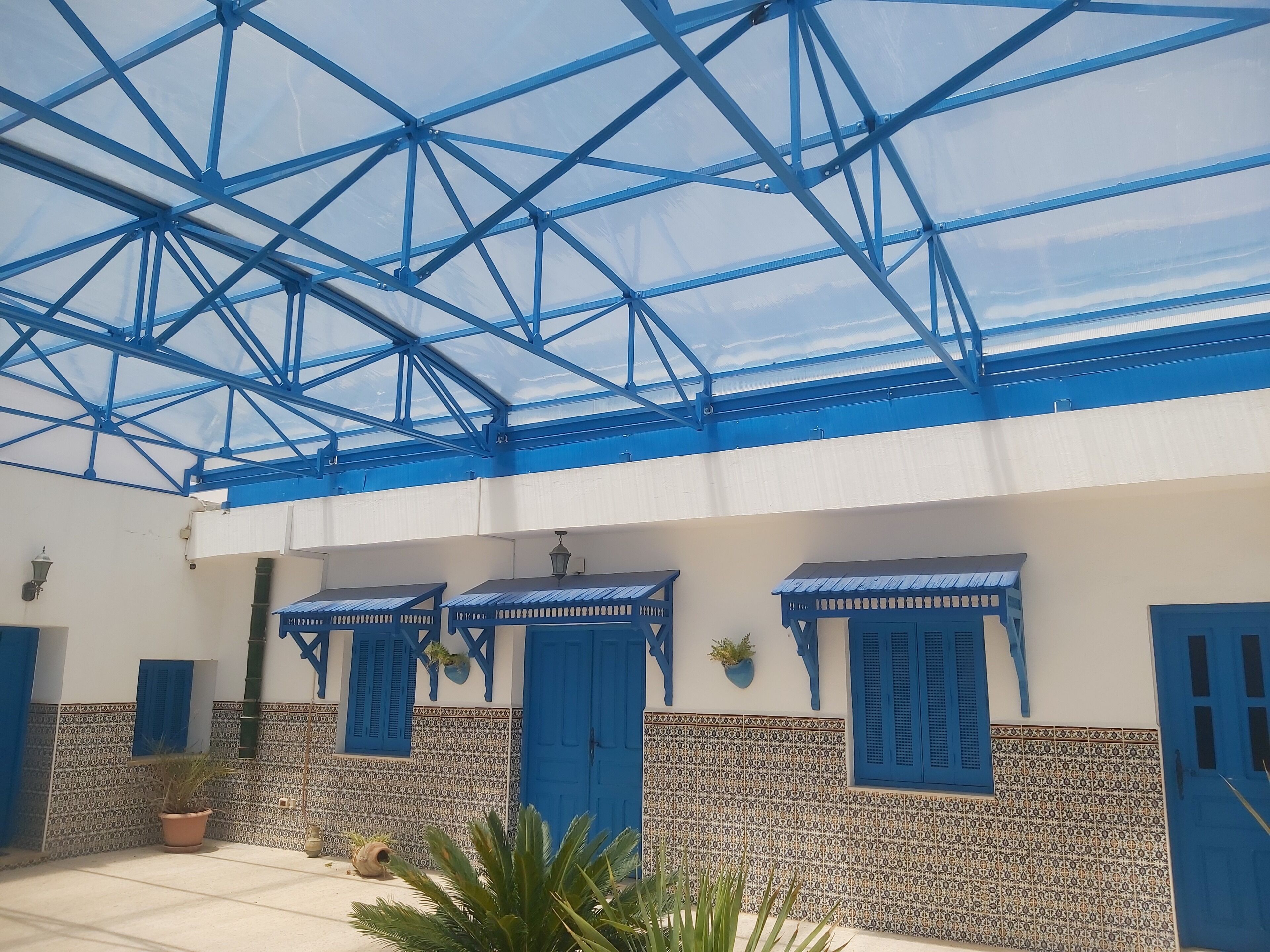 A covered outdoor area with blue metal beams and a glass roof, potted plants, and blue doors with shutters.