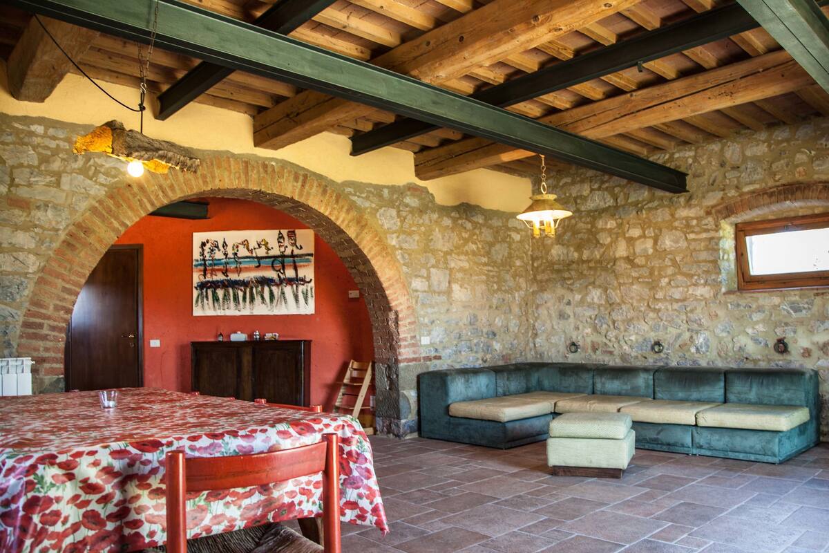 A rustic room with a stone wall, wooden ceiling beams, a dining table with a floral tablecloth, a green sofa, and a wooden door.