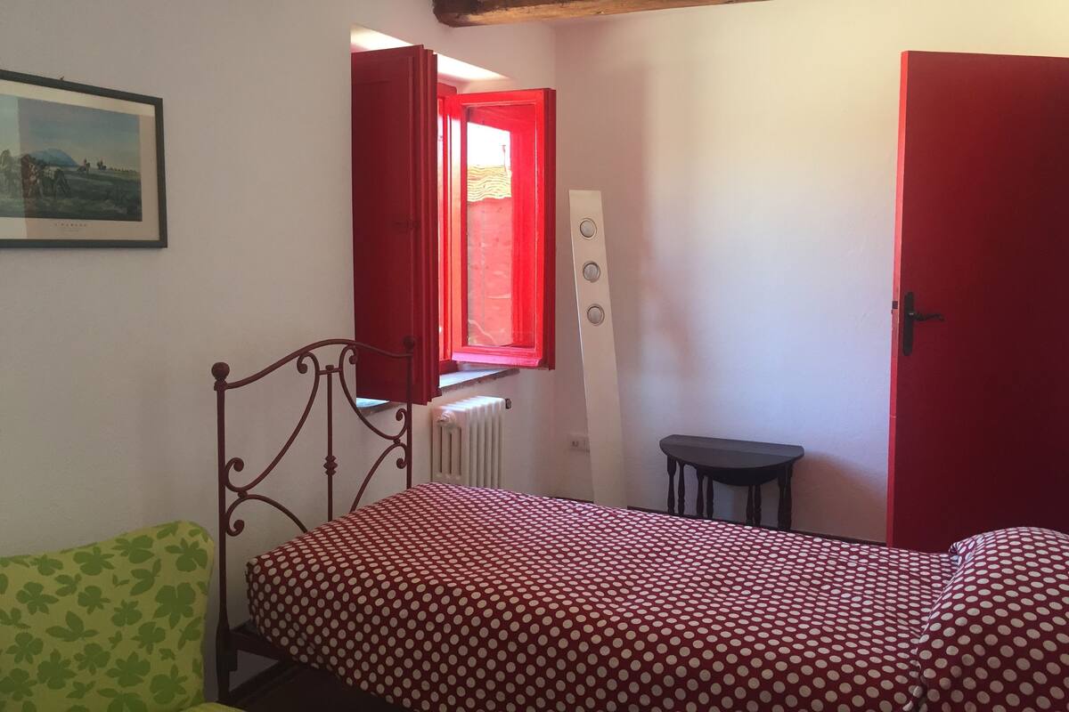 A bedroom with a bed, a chair, a radiator, a window with red curtains, and a door.