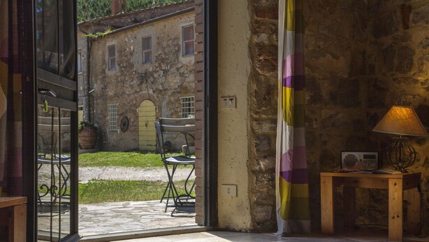 An open door leading to an outdoor patio with a table and chairs, a stone building in the background, and a lamp on a small table inside. - Agriturismo La Valentina Nuova (Orbetello)