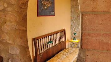Una stanza con parete in pietra, un letto con testiera in legno, un tavolino con una lampada e un quadro appeso al muro.