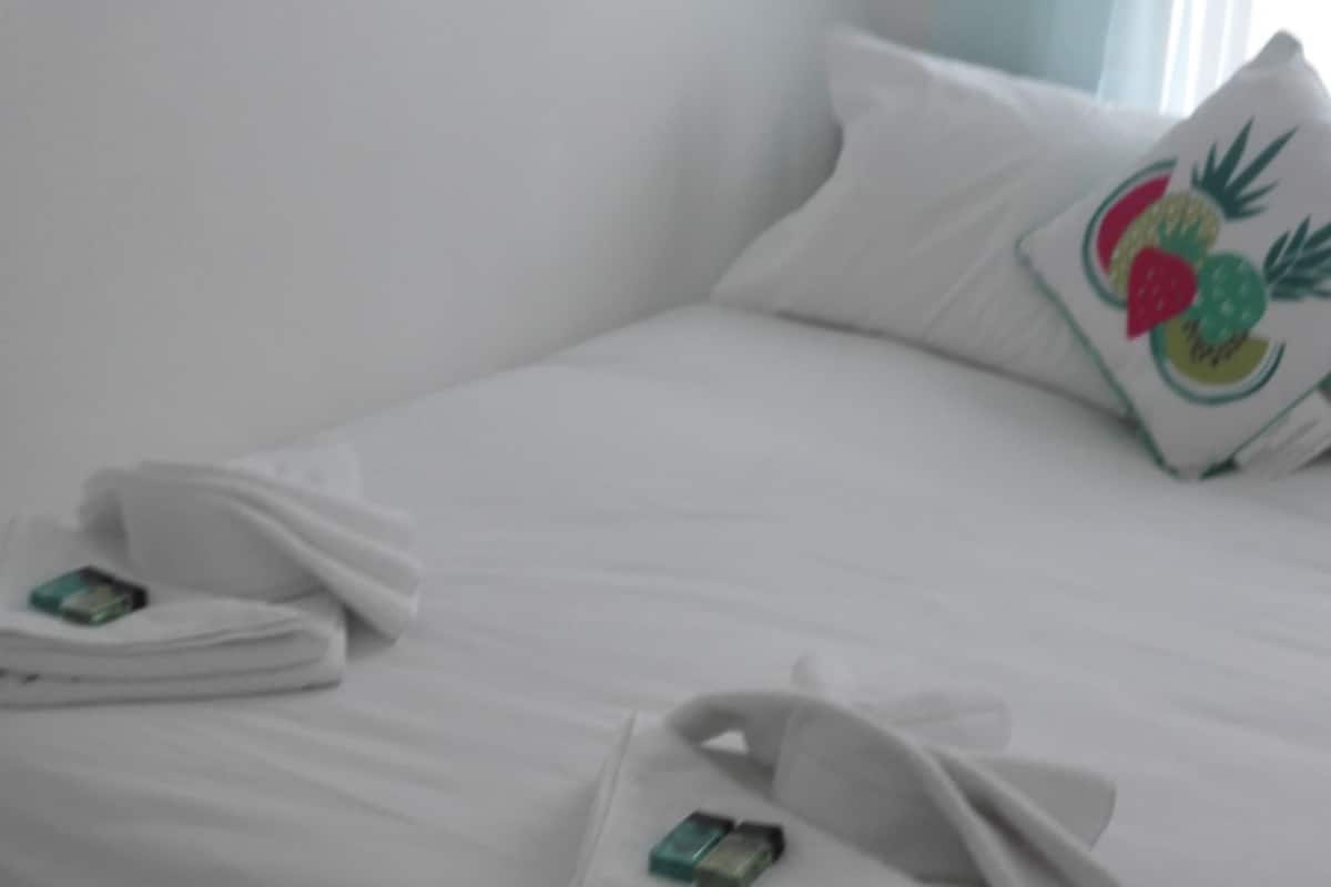 A neatly made bed with towels folded on it, a pillow with a fruit design, and a wooden floor.