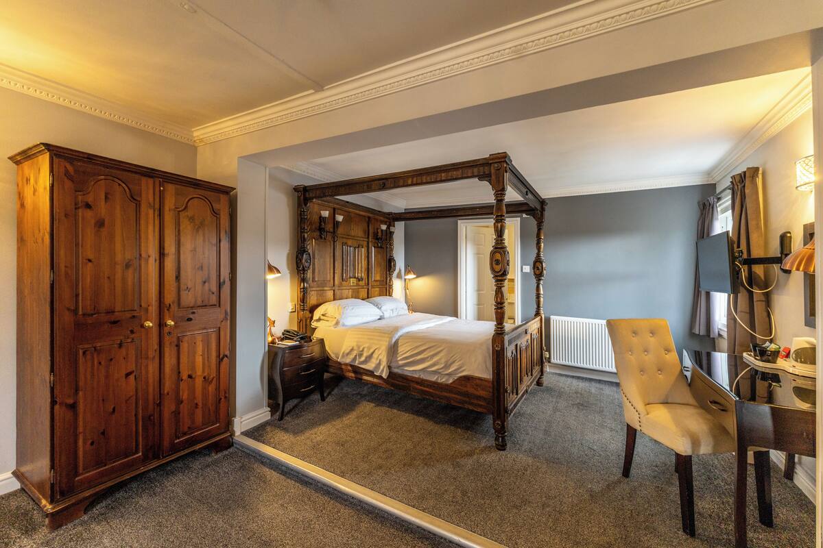 A four-poster bed with a wooden frame, a nightstand, a chair, and a wardrobe in a room with a carpeted floor and a radiator.