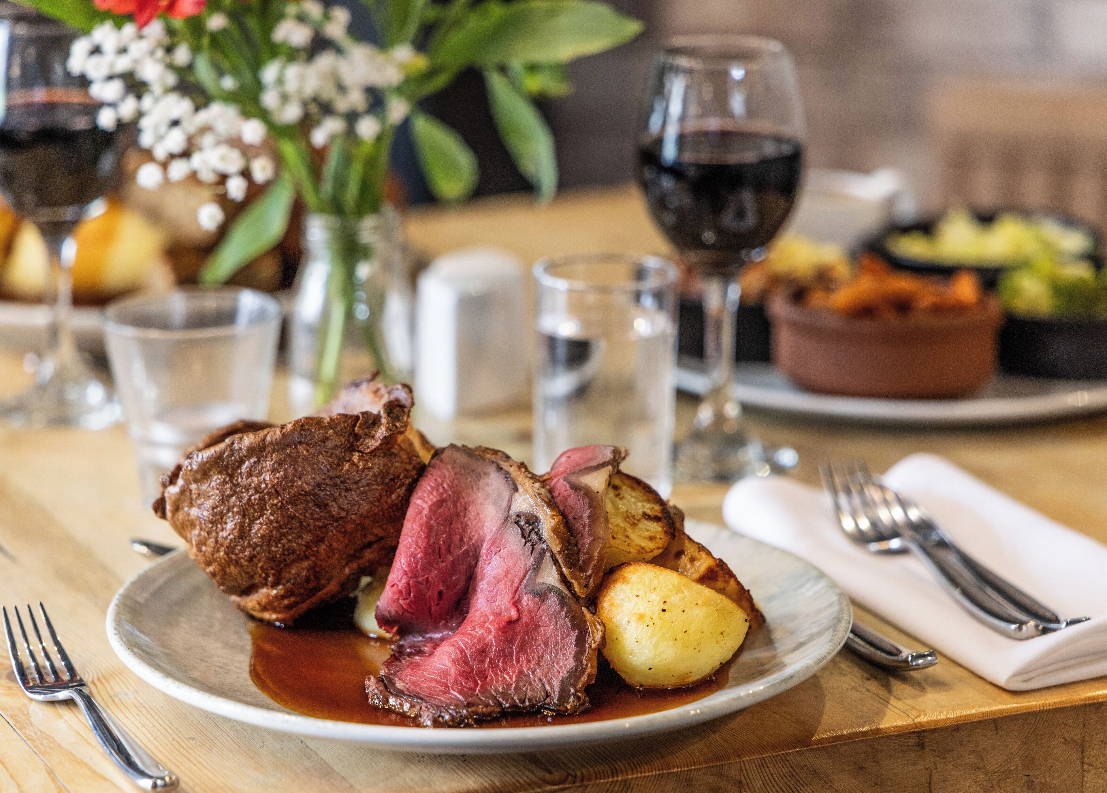 A plate of steak with roasted potatoes and gravy, a glass of red wine, and a vase of flowers on a wooden table.