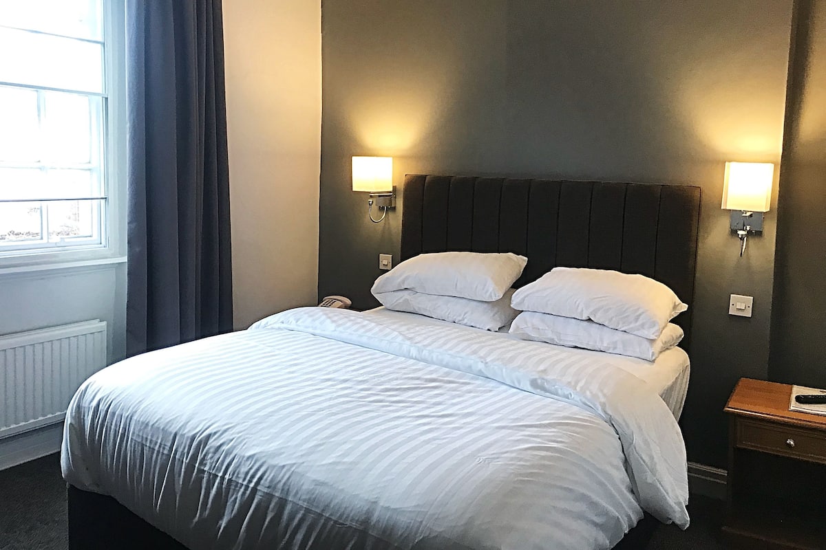 A hotel room with a bed, two bedside lamps, a nightstand, and a window with curtains.