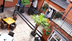 Een terras buiten met tafels en stoelen, een klein tuintje met planten en een bakstenen gebouw met ramen en een deur.