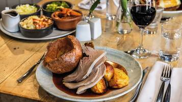A plate of roasted meat and potatoes on a wooden table with a glass of red wine, salt and pepper shakers, and small bowls of side dishes.