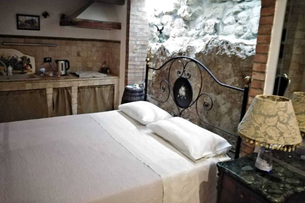 Una cama con sábanas blancas, una pared de piedra, una mesita con una lámpara y una zona de cocina con hervidor y otros objetos.