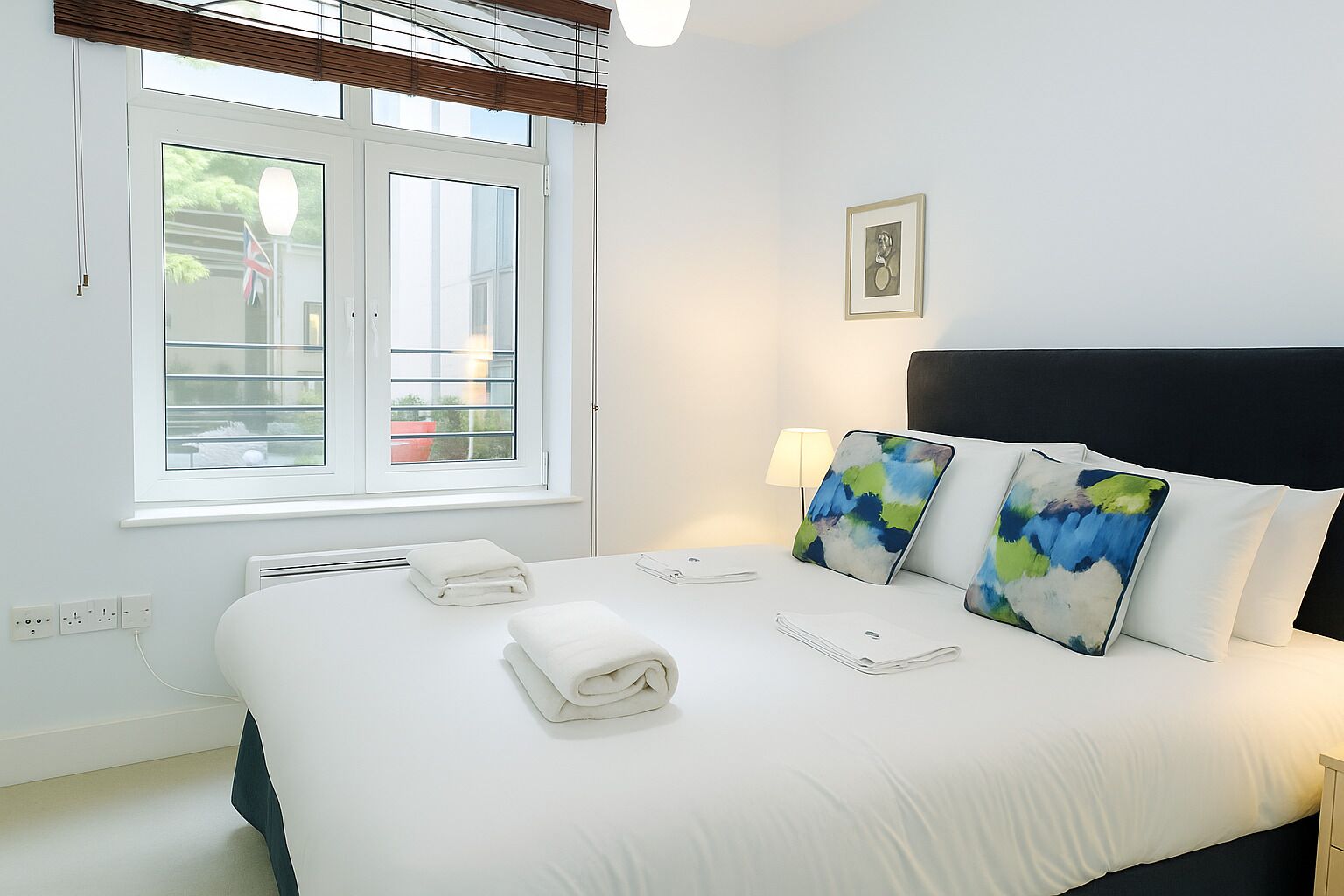 A hotel room with a large bed, a window with blinds, a bedside lamp, and a framed picture on the wall.