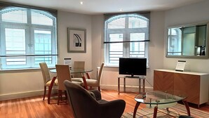 Ein modernes Wohnzimmer mit Glastisch, bequemen Sitzmöglichkeiten, einem Flachbildfernseher und großen Fenstern mit Jalousien.