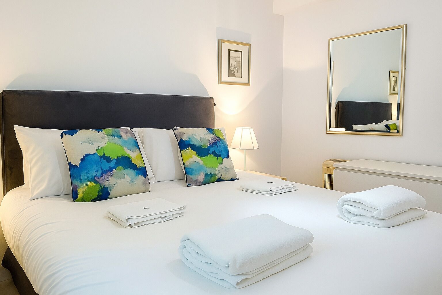 A hotel room with a large bed, two colorful pillows, a bedside lamp, a mirror, and neatly folded towels.