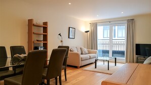 Ein modernes Wohnzimmer mit einer Couch, Sesseln, einem Couchtisch und einem Fernseher. Es gibt einen Balkon mit Geländer und ein Fenster mit Vorhängen.