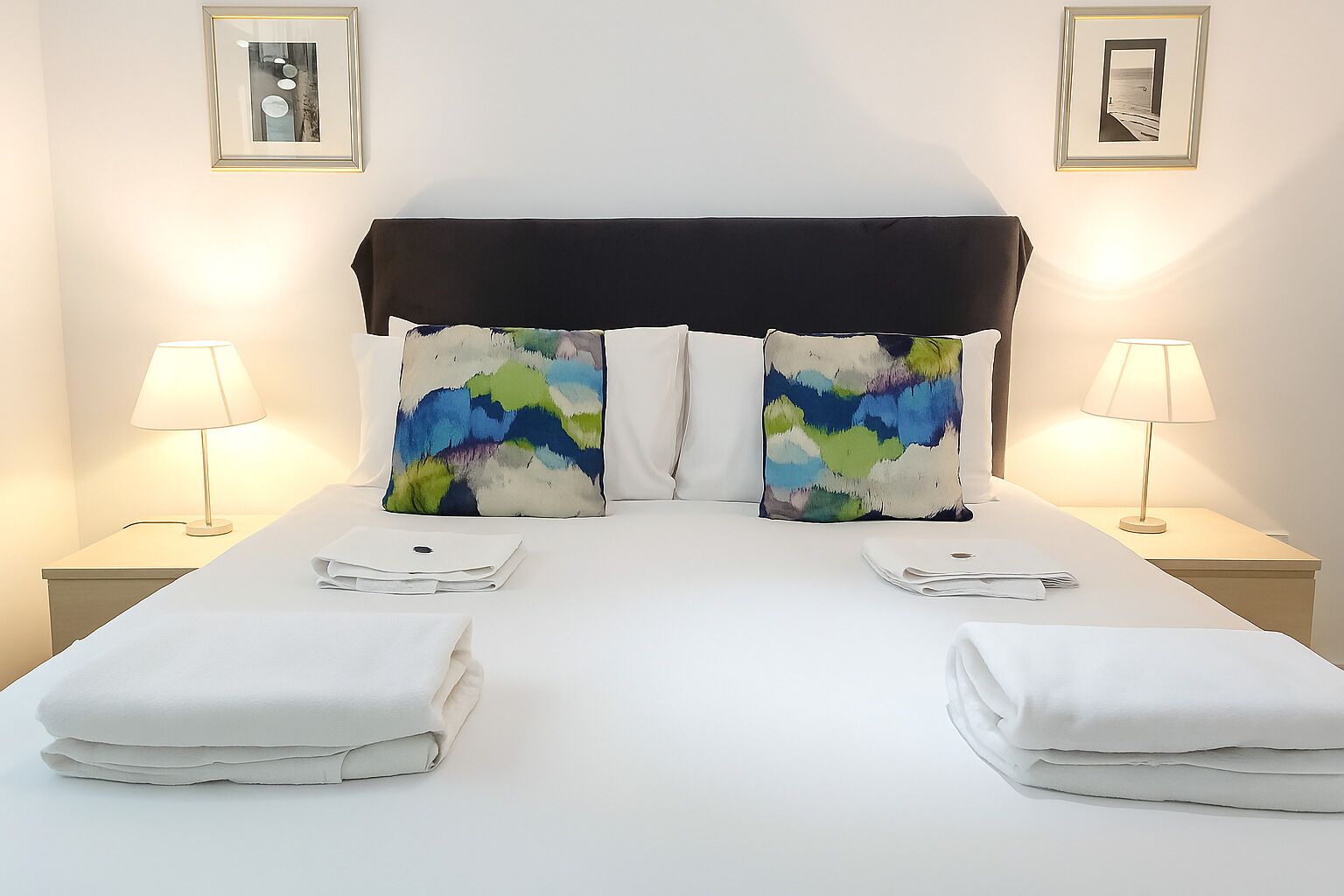 Cama bem arrumada com duas almofadas, duas luminárias de cabeceira e duas toalhas dobradas sobre um lençol branco.