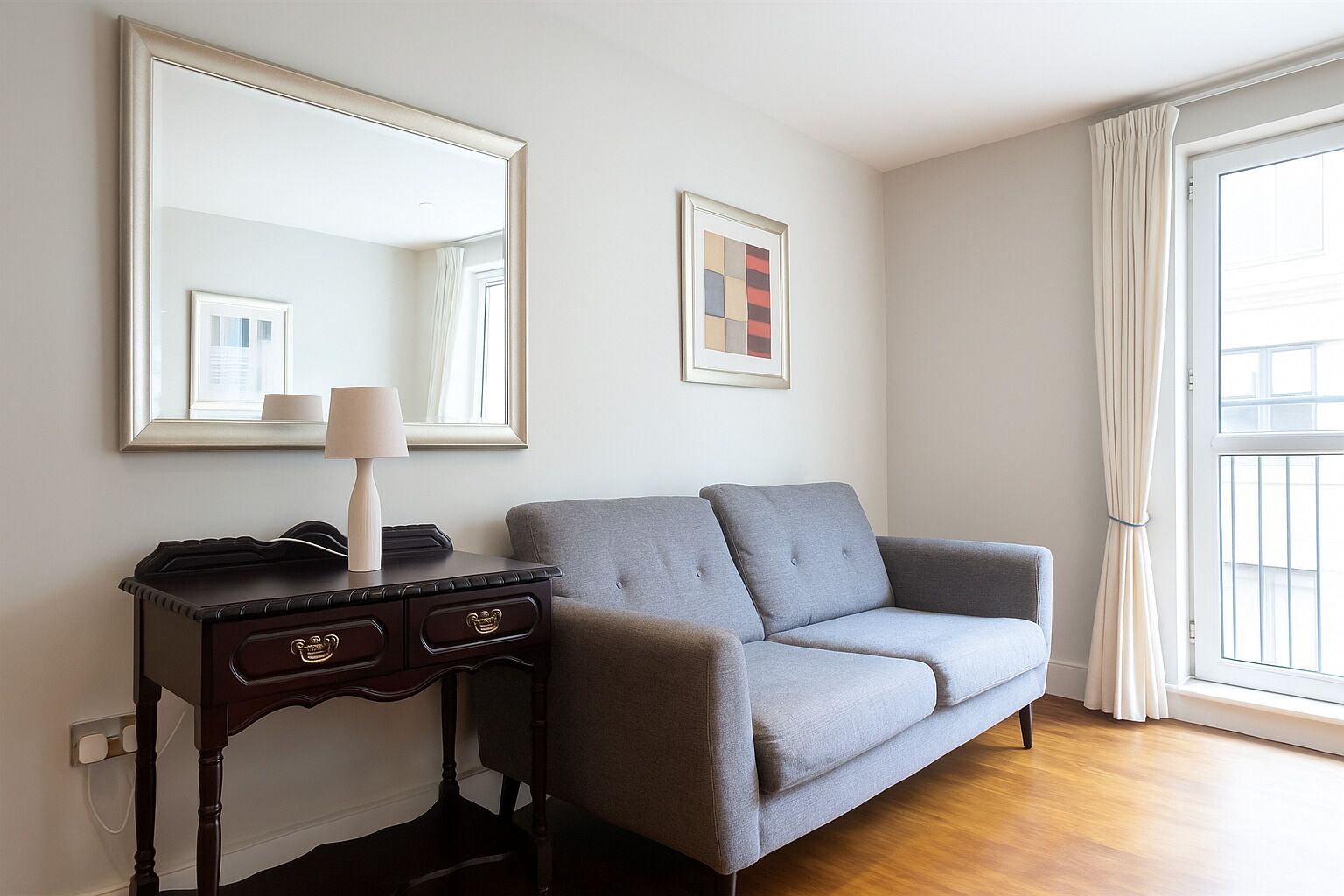 A room with a grey sofa, a dark wooden side table with a lamp, a large mirror, and a framed picture on the wall.