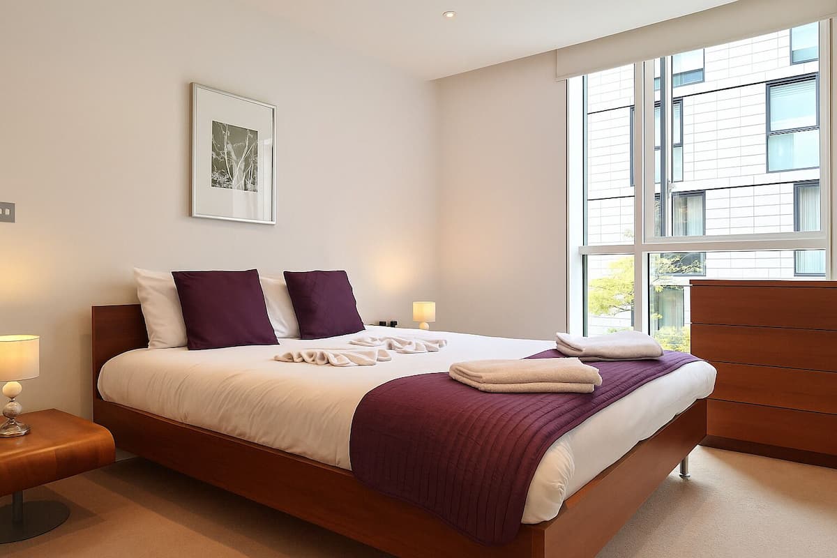 A modern bedroom with a large bed, two bedside lamps, a wooden dresser, and a large window with a view of buildings.