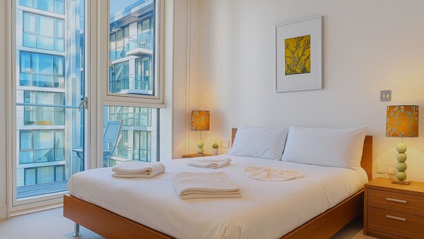 A modern hotel room with a large bed, two bedside tables, a window with a view of a building, and two wall-mounted lamps.