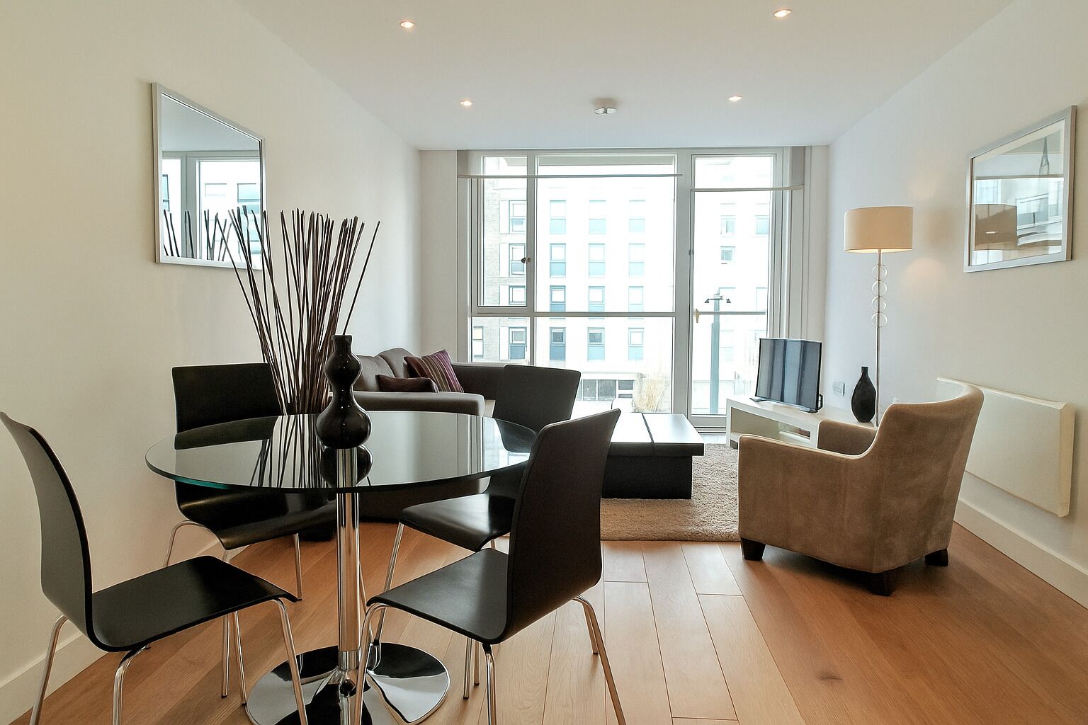 Een moderne woonkamer met een ronde glazen tafel, zwarte stoelen, een beige bank en een televisie.