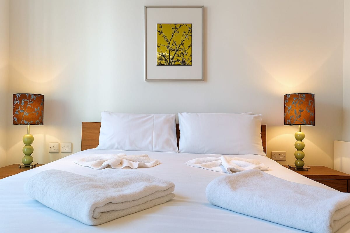 A hotel room with a neatly made bed, two pillows, two lamps, and a framed artwork on the wall.