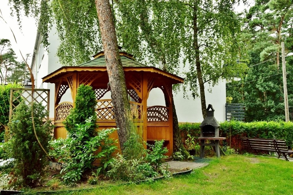 A wooden gazebo with a lattice design, surrounded by lush greenery and a stone oven.
