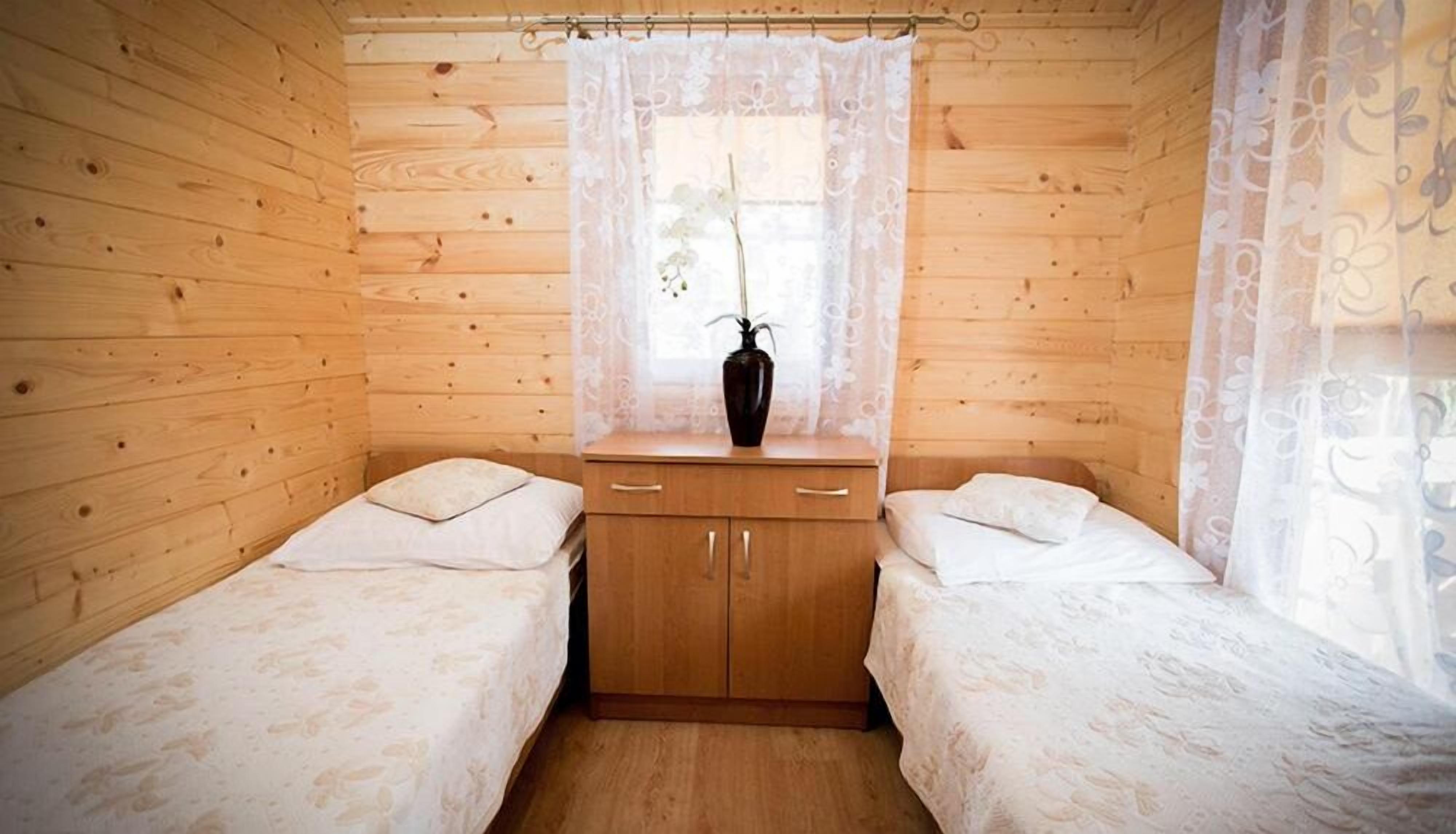 Ein kleines Zimmer in einer Holzhütte mit zwei Einzelbetten, einem Holzkleiderschrank und einem Fenster mit Spitzenvorhängen.