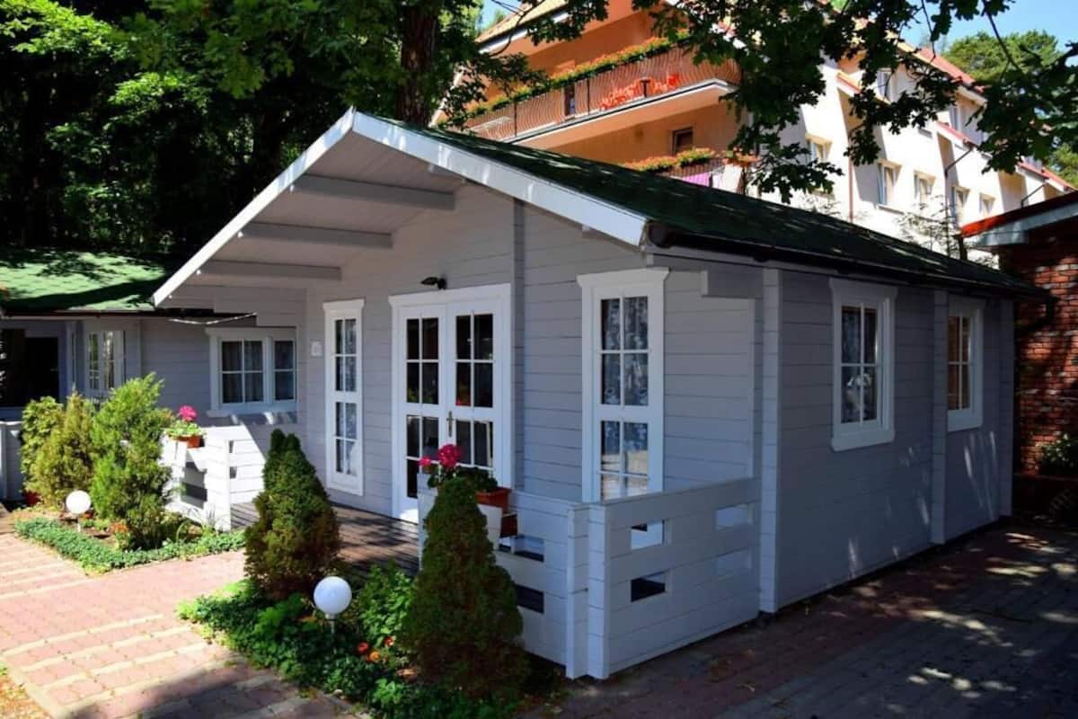 Une maison à deux étages, à l’extérieur blanc et au toit vert, entourée d’un chemin en briques et de petits arbres.