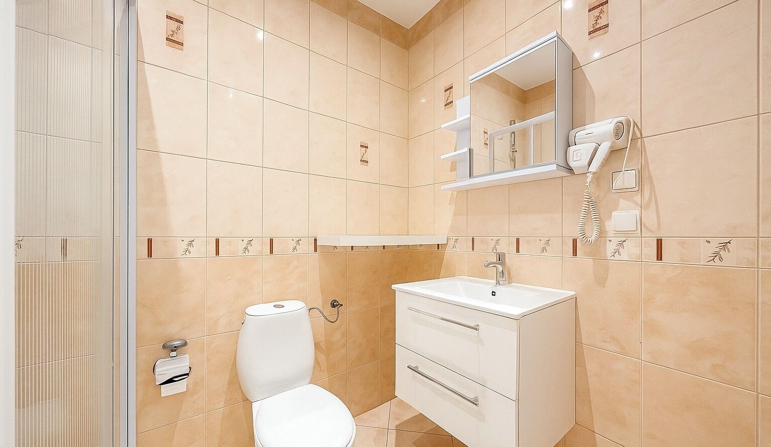 A bathroom with beige tiles, a toilet, a sink, a mirror, and a hairdryer.