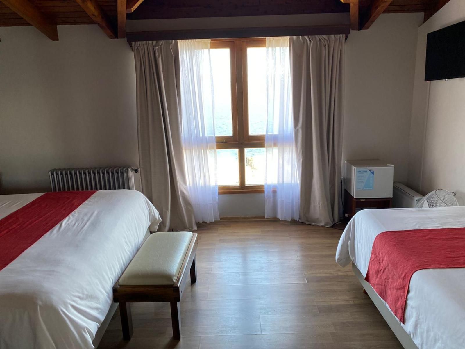 A hotel room with two beds, a bench, a radiator, a small TV, and a window with curtains.