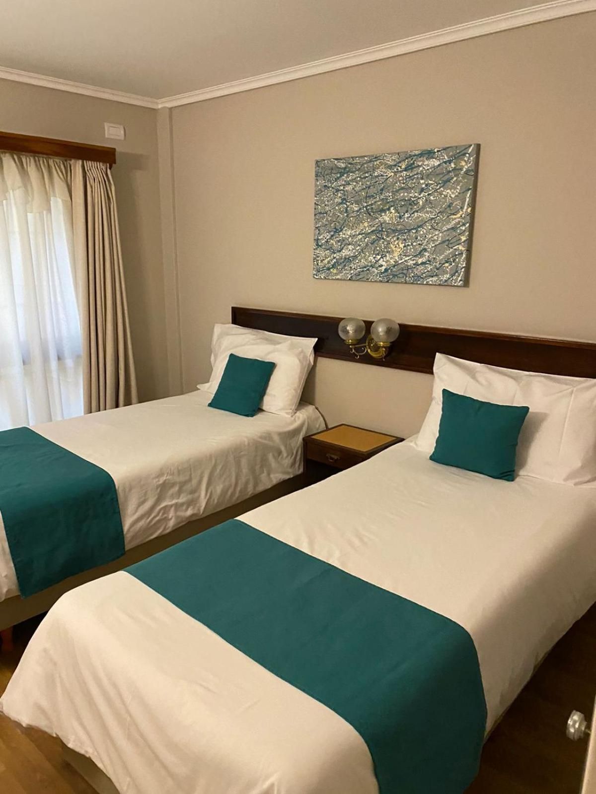 A hotel room with two beds, each with white bedding and teal accents, a nightstand with lamps, and a painting on the wall.
