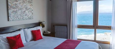 Quarto de hotel com uma cama grande, roupa de cama vermelha e branca, vista para o mar e teto com vigas à vista.