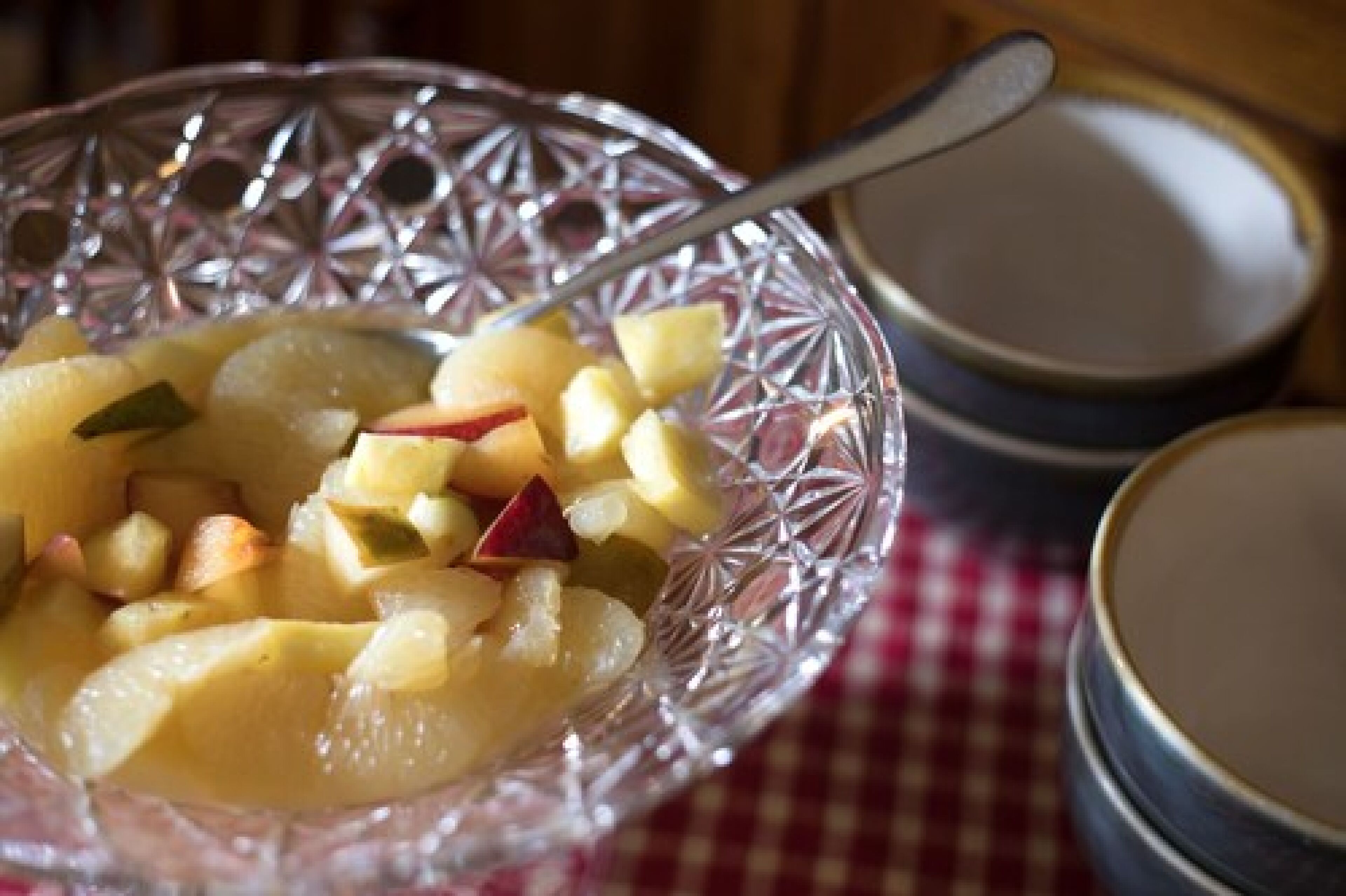 A clear glass bowl with diced apples and a spoon, placed on a checkered tablecloth.