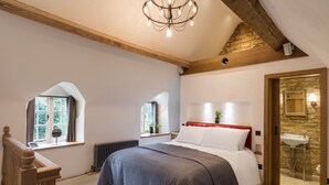 Ein Schlafzimmer mit einem großen Bett, Holzbalken an der Decke, einer Kristallleuchtre, einem Fenster mit Vorhängen, einem Heizkörper und einer Steinwand.