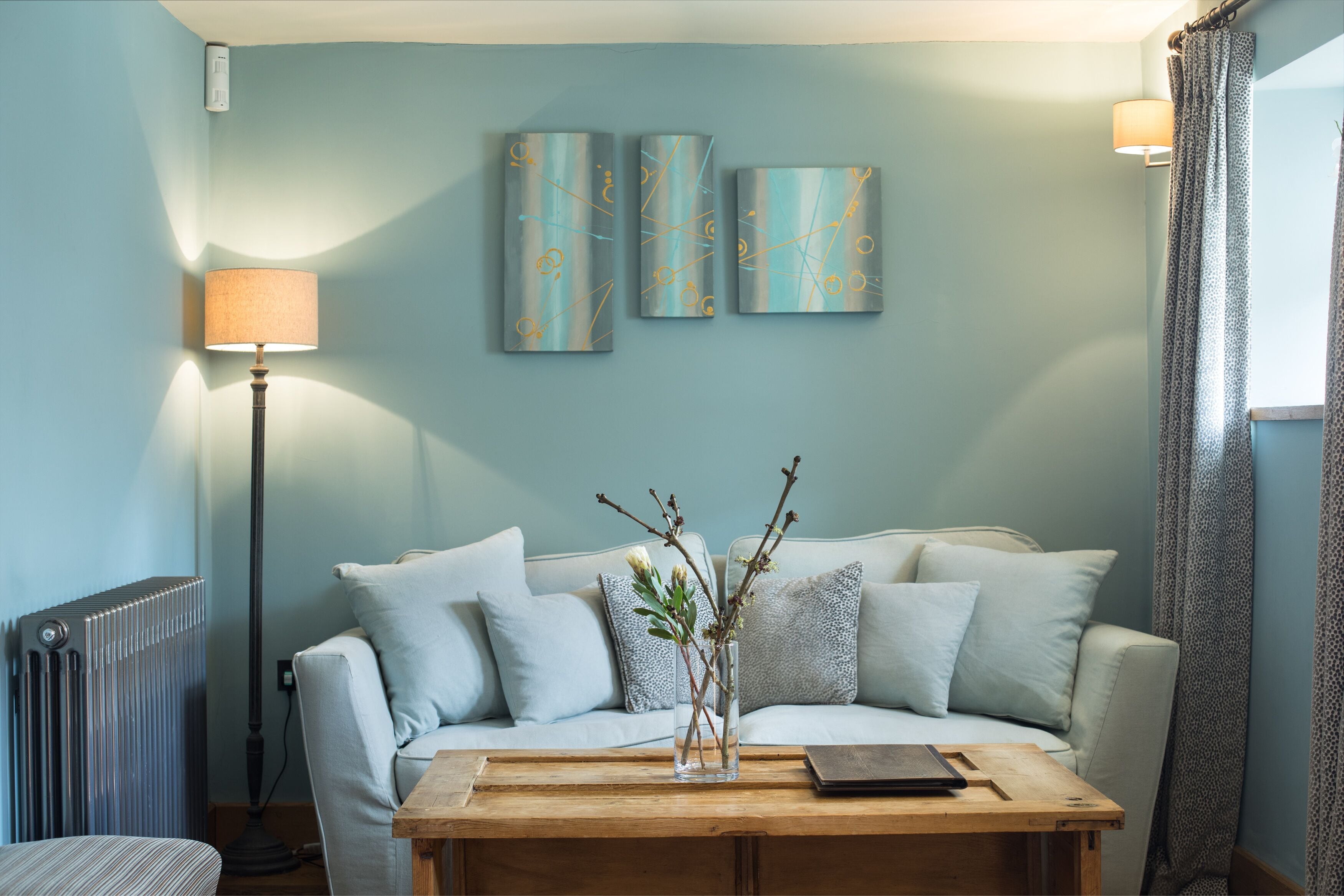 A living room with a sofa, a coffee table, and a lamp. There are three wall-mounted artworks above the sofa.