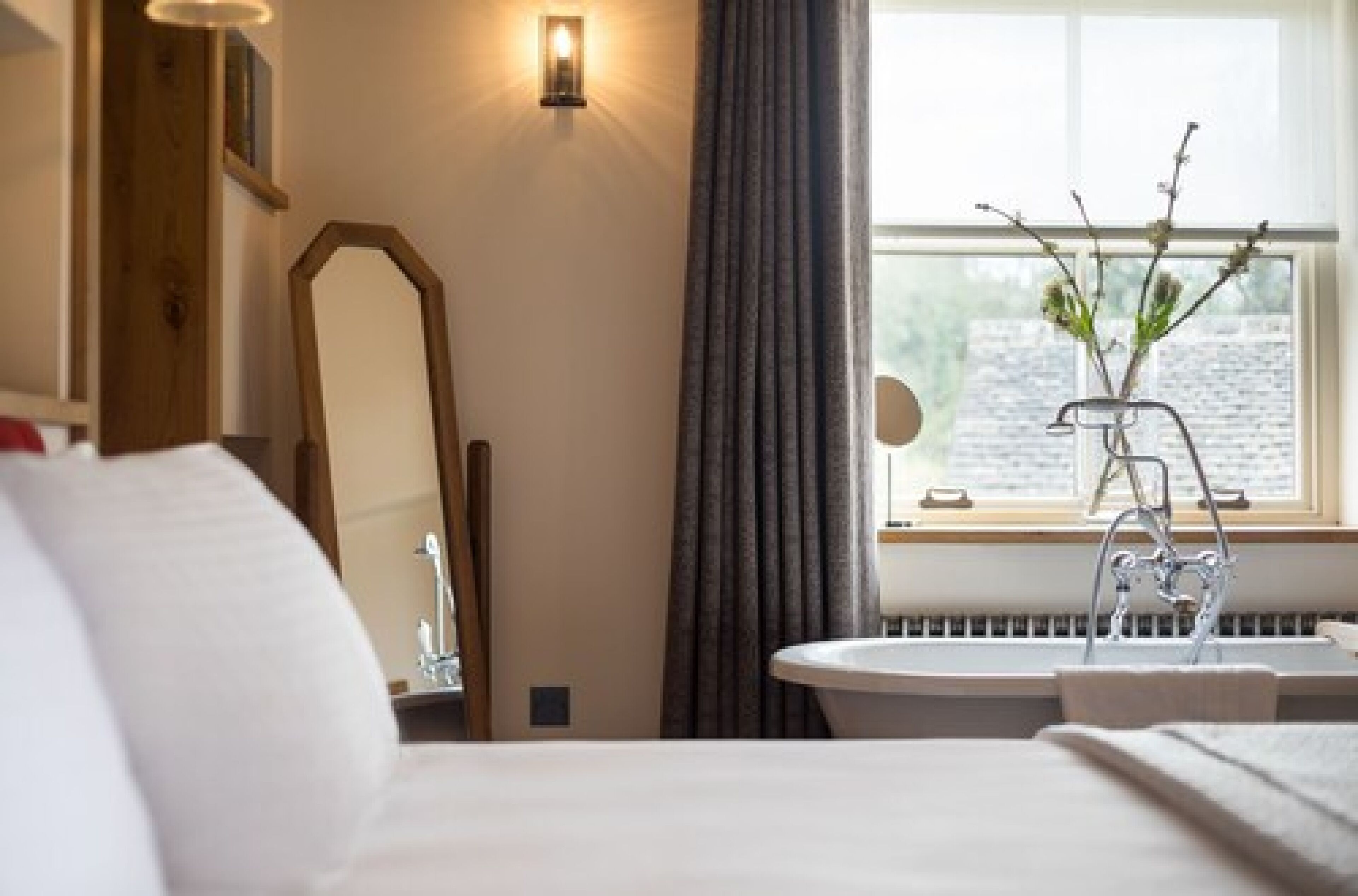 A hotel room with a bed, a round mirror, a window with curtains, a radiator, and a vase with branches.