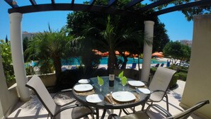 A glass-top table with four chairs, a bottle of wine, and a glass of water, set against a backdrop of a pool and palm trees. - Villa Hesperides Gardens (Paphos)