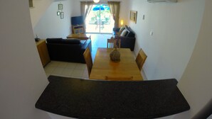 A modern kitchen with a wooden dining table, a black countertop, and a view into the living area with a sofa and a TV. - Villa Hesperides Gardens (Paphos)