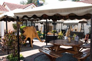 A patio area with a wooden table and benches, surrounded by umbrellas and potted plants. - Mpofu Guest House (Harare)