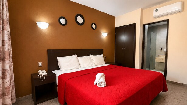 Habitación de hotel con una cama, una mesita de noche, un teléfono, una colcha roja, una toalla y un aire acondicionado de pared.