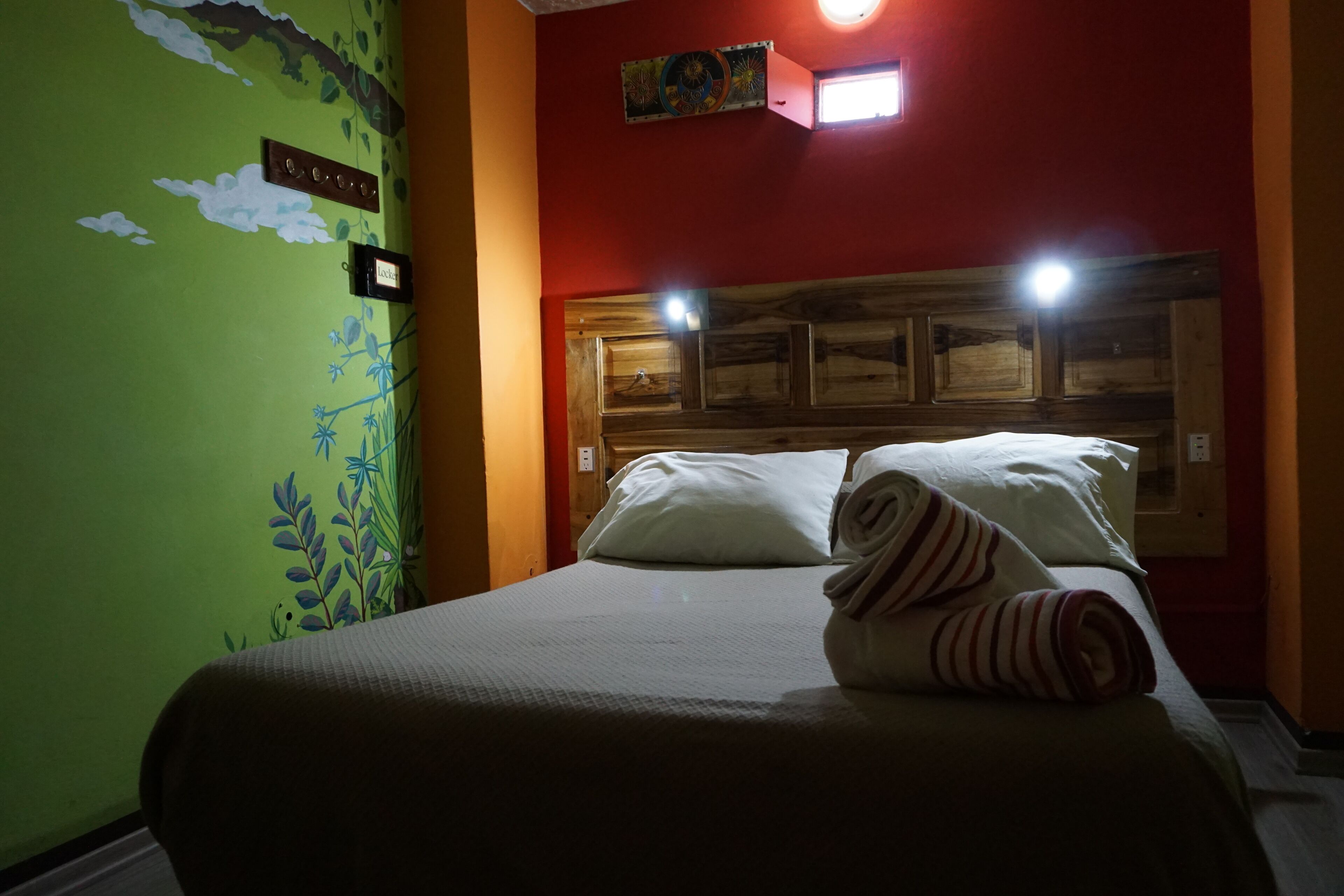 A hotel room with a bed, a headboard with lights, a green wall with a floral mural, and a small painting above the headboard.
