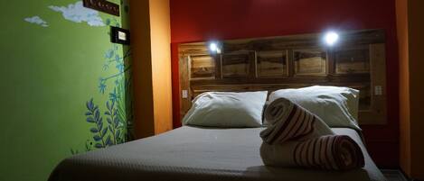 A hotel room with a bed, a headboard with lights, a green wall with a floral mural, and a small painting above the headboard.