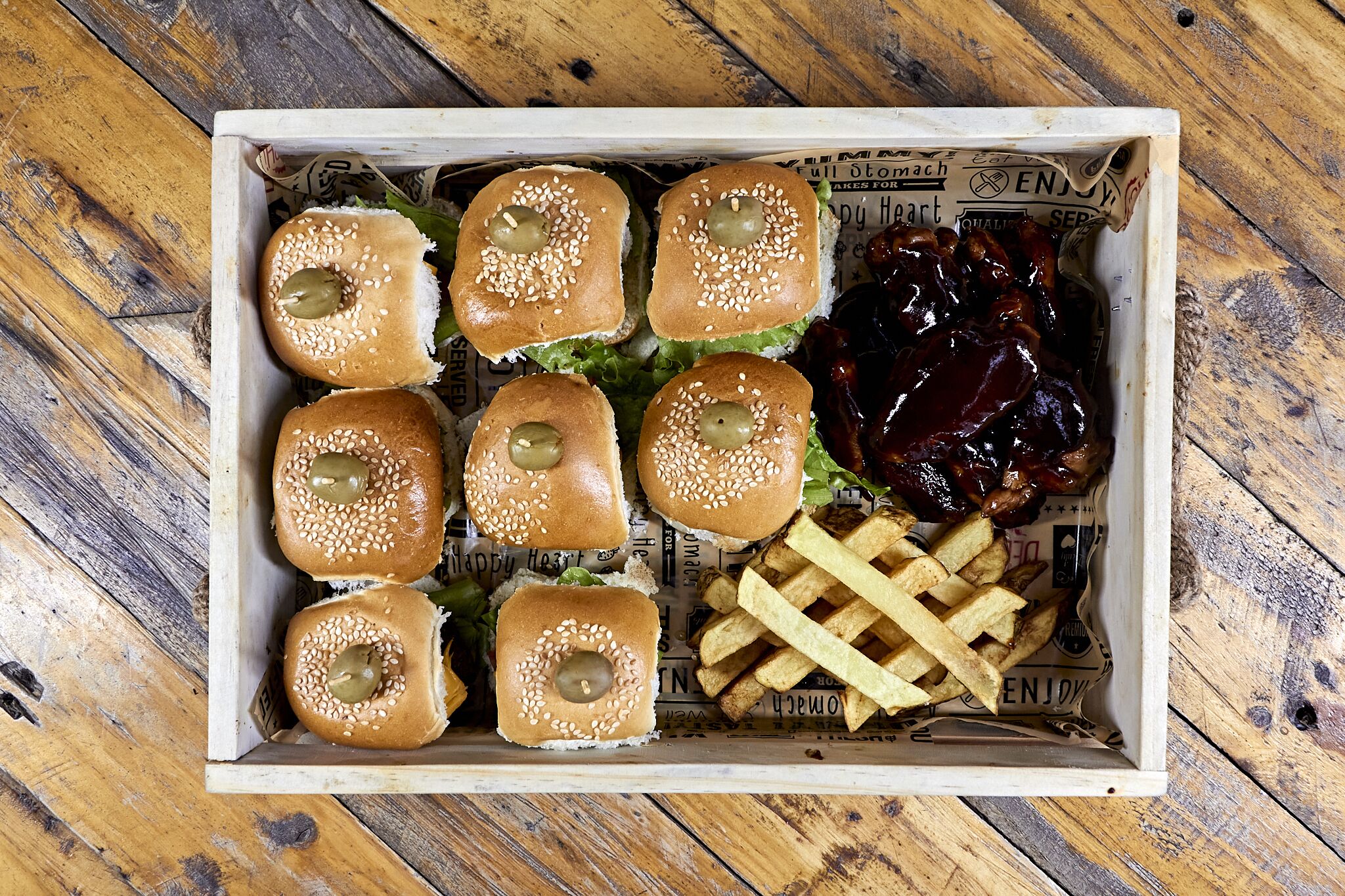 Una caja de madera que contiene seis bollos de semillas de sésamo con aceitunas verdes, una porción de papas fritas y salsa de arándanos.