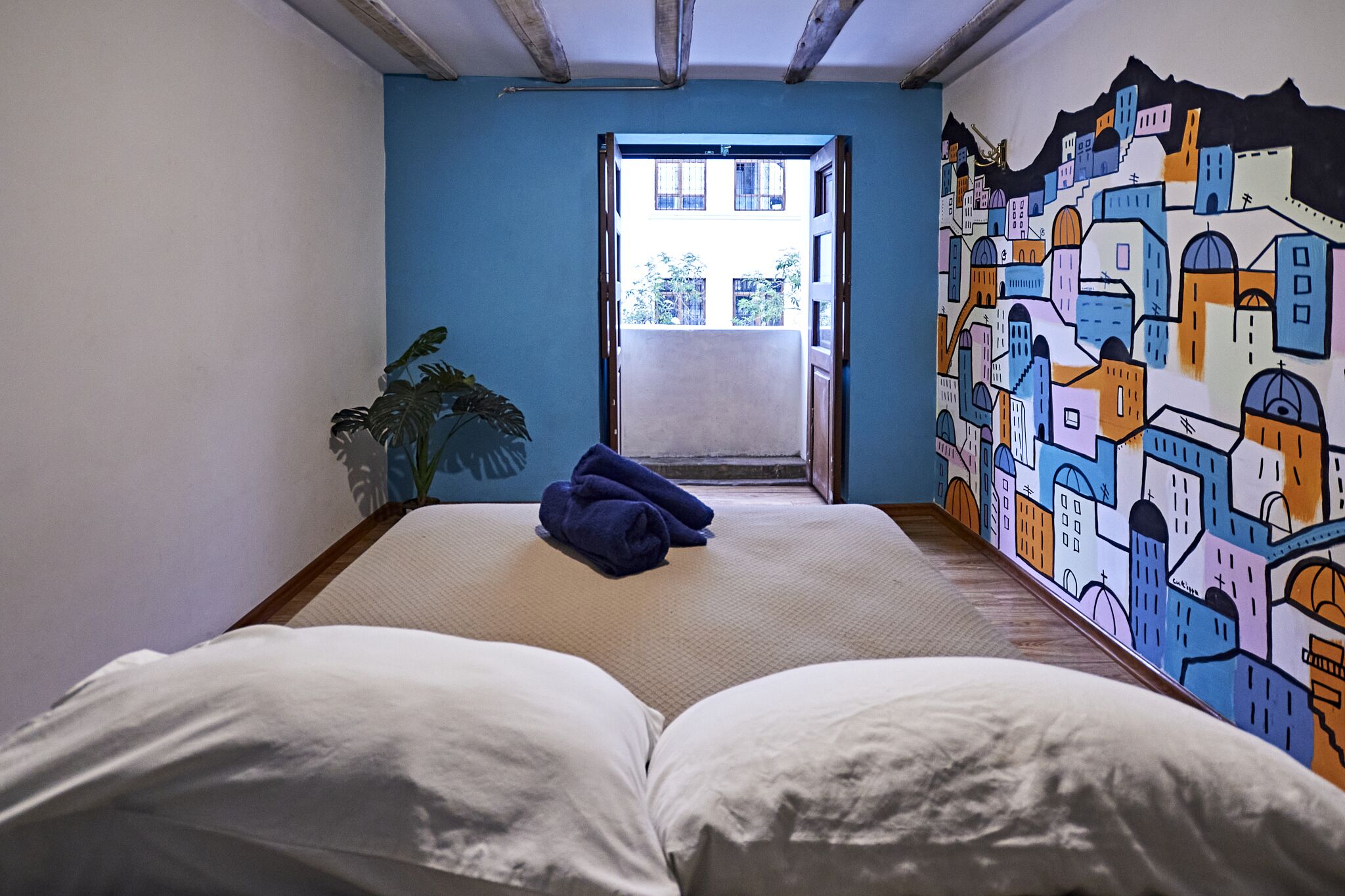 A room with a bed, a blue wall with a colorful mural, a potted plant, and a view of a building through an open door.