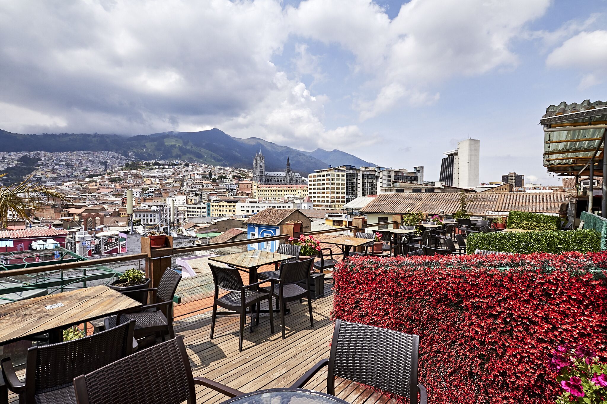 A rooftop terrace with wooden tables and chairs, a flower bed, and a cityscape with mountains in the background.