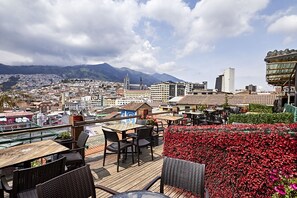 A rooftop terrace with wooden tables and chairs, a flower bed, and a cityscape with mountains in the background.
