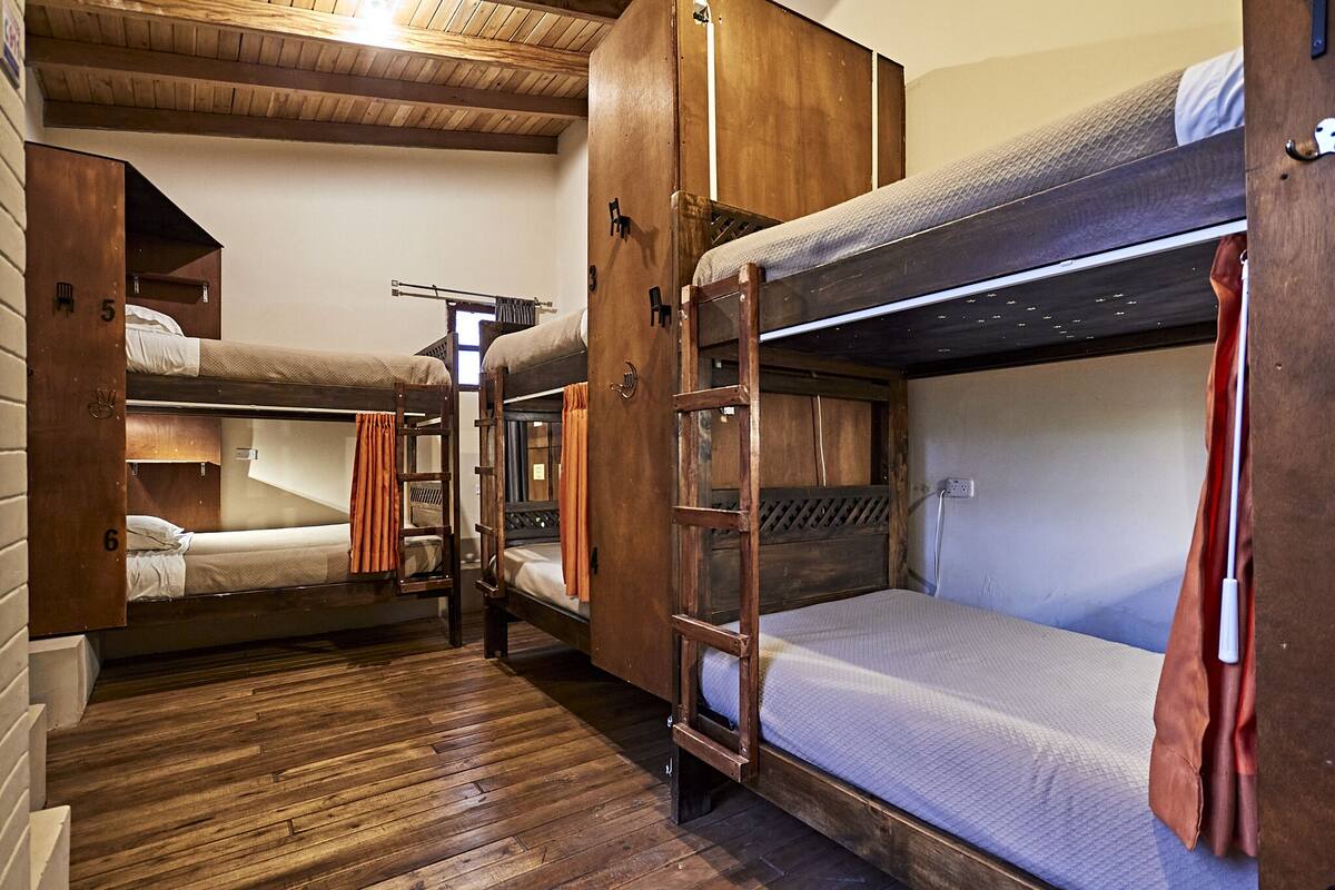 A dormitory-style room with bunk beds, wooden floors, and a ladder.