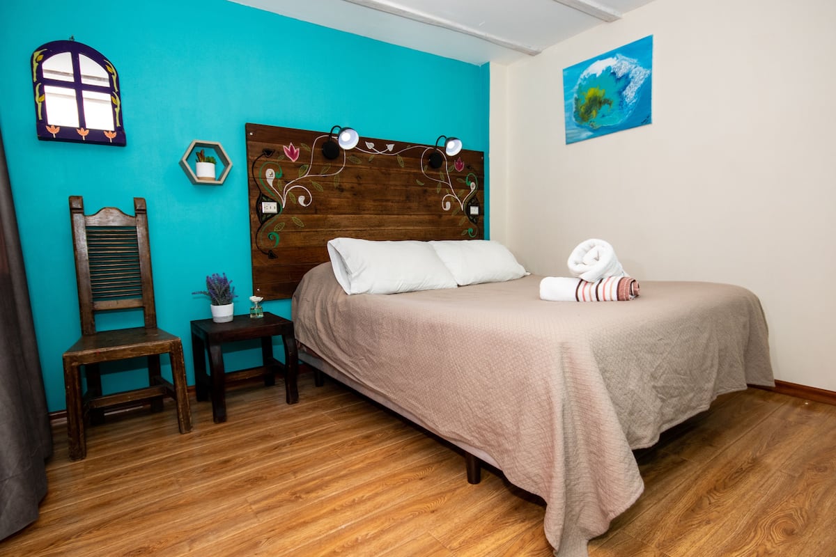 A bedroom with a wooden headboard, a bed with a beige bedspread, a small bedside table with a plant, and a chair.