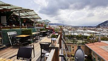 An outdoor terrace with wooden tables and chairs, a covered seating area, and a view of the cityscape.