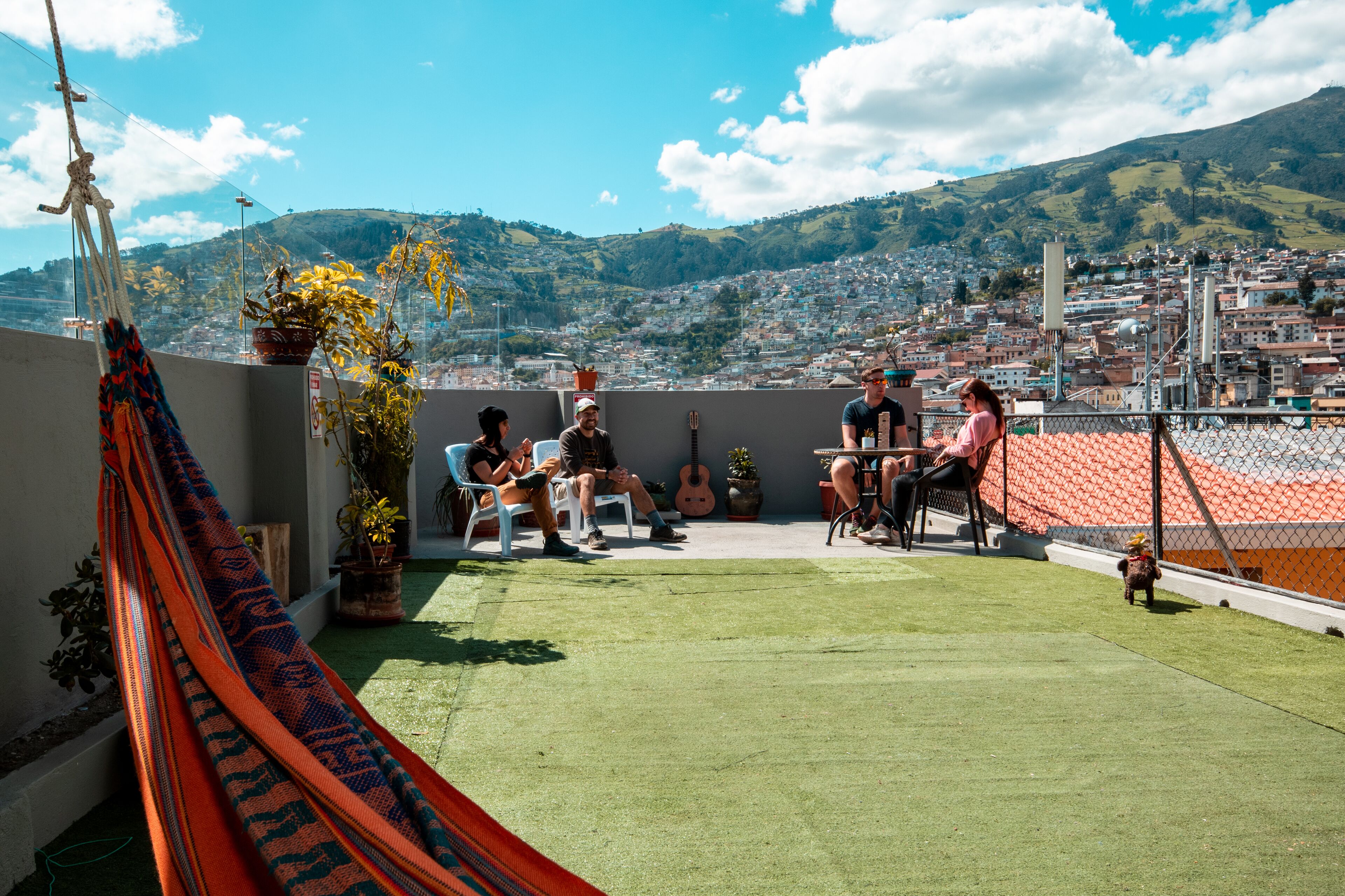 A rooftop terrace with a hammock, potted plants, and a view of a cityscape and hills.