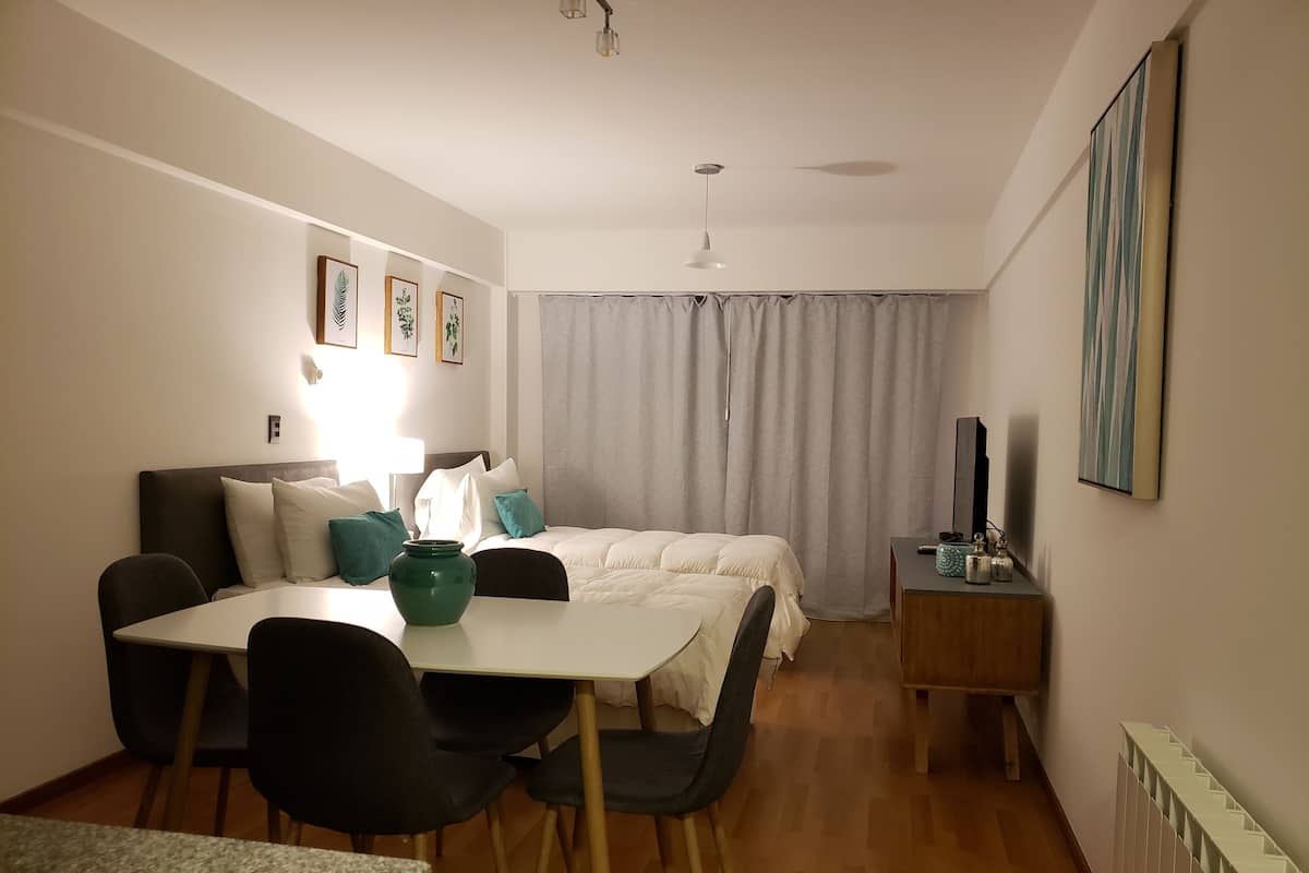 Hotellihuone, jossa on sänky, työpöytä, tuoleja, pöytä, televisio ja patteri.