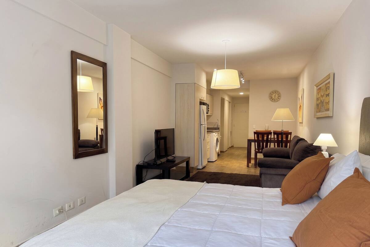 Hotellihuone, jossa on sänky, työpöytä tietokoneella, tuoli, ruokapöytä ja keittiöalue.